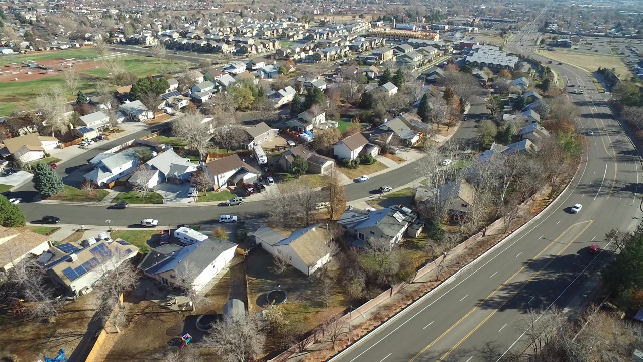High aerial view over Westminster, Colorado homes, leafless trees, lawns, a curving divided road, and distant fields with no visible people.