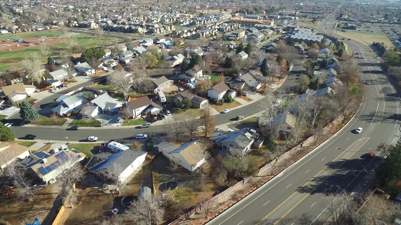 Aerial frame of Westminster, Colorado rooftops, backyards, mature trees, parked vehicles, and a broad road running along the right side with no visible people.