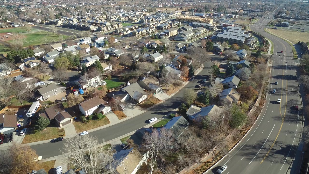 A high aerial frame over Westminster, Colorado shows residential roofs, curved side streets, leafless trees, sports fields in the distance, and light traffic on a multi-lane road with no visible people.