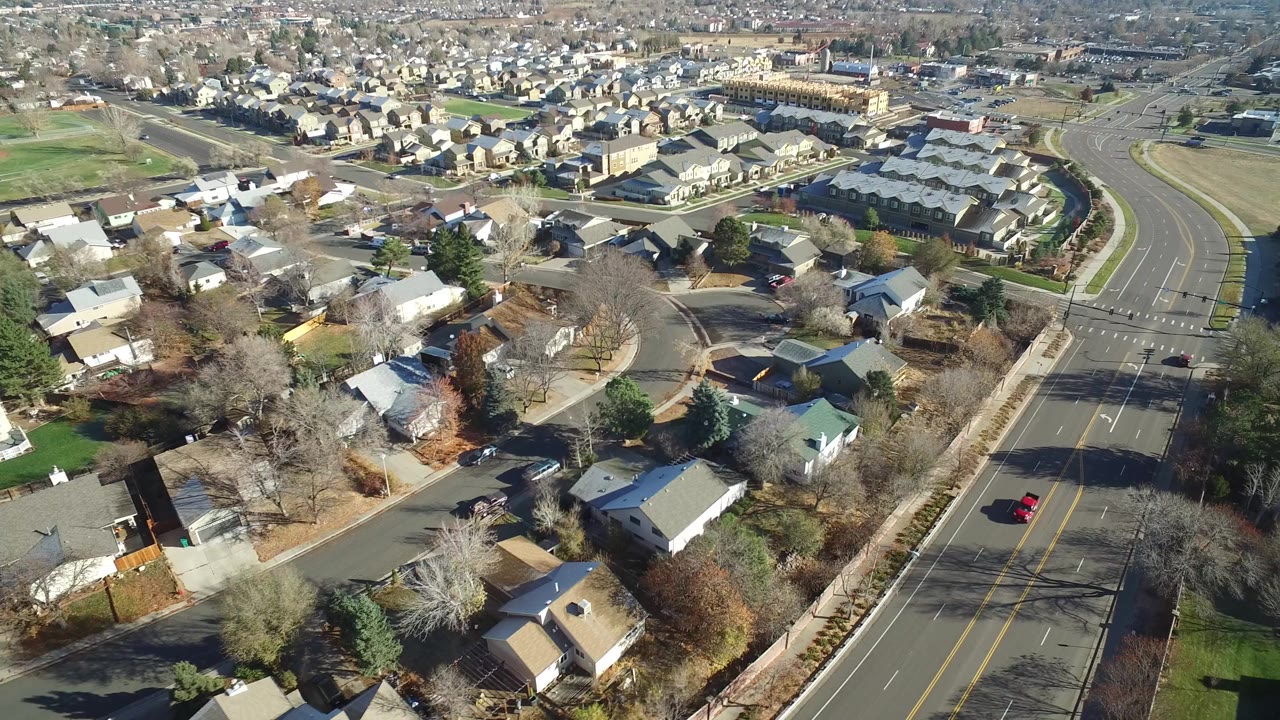 Aerial view of suburban homes, trees, and a wide road in bright sun.