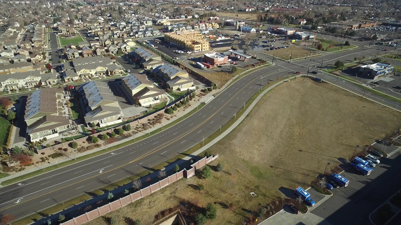 Opening aerial frame over Westminster, Colorado showing a dry grass lot beside multi-lane roads, townhomes, parked cars, and commercial buildings, with no visible people.