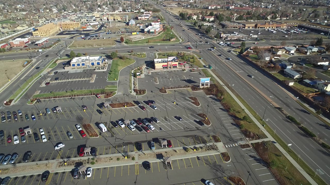 The second frame keeps the Westminster aerial angle on the retail parking area, marked stalls, broad roadway, and low residential neighborhood beyond, with no visible people.