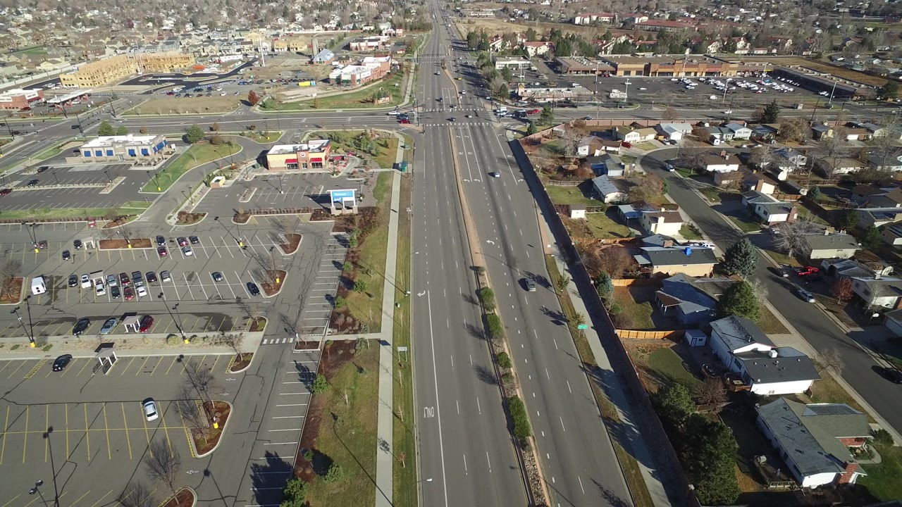 High aerial frame in Westminster, Colorado with a broad road centered near parking lots, houses, lawns, sidewalks, scattered cars, and no visible people.