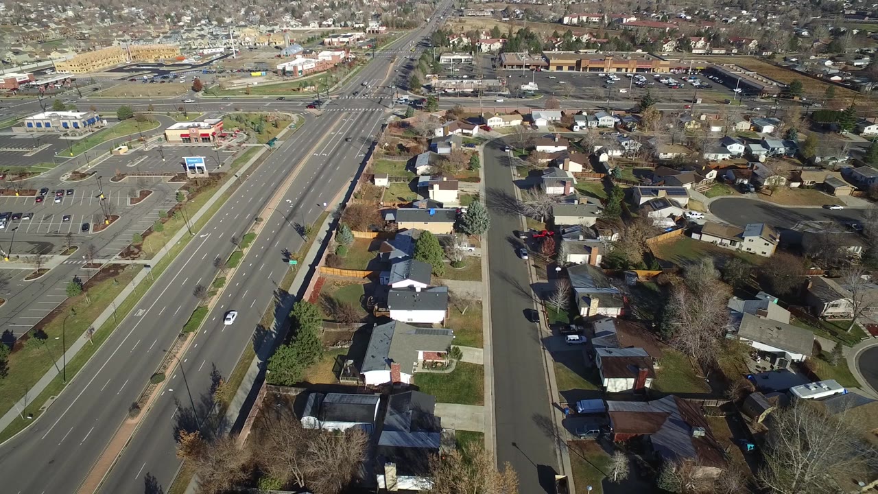 Aerial frame in Westminster, Colorado with rooftops and lawns in the foreground, a long multi-lane road at left, scattered cars, and no visible people.