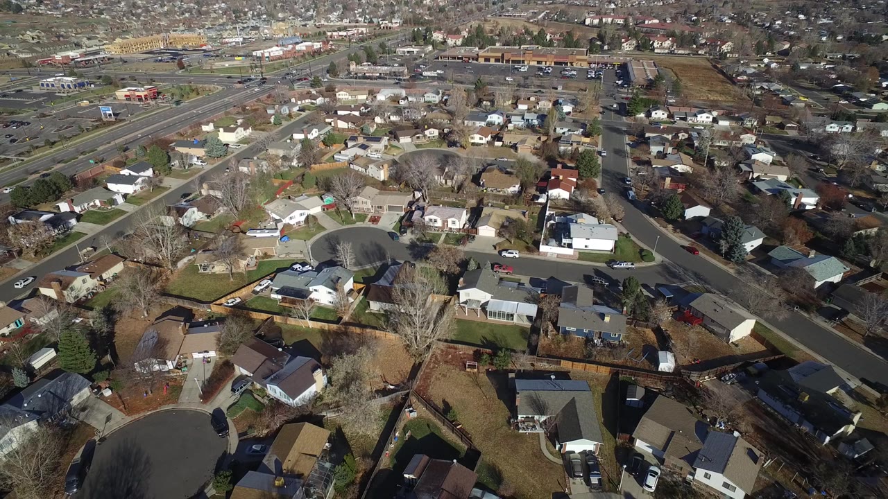 Aerial frame over Westminster, Colorado showing a curving neighborhood road, closely spaced homes, fenced backyards, leafless trees, and no clearly visible people.