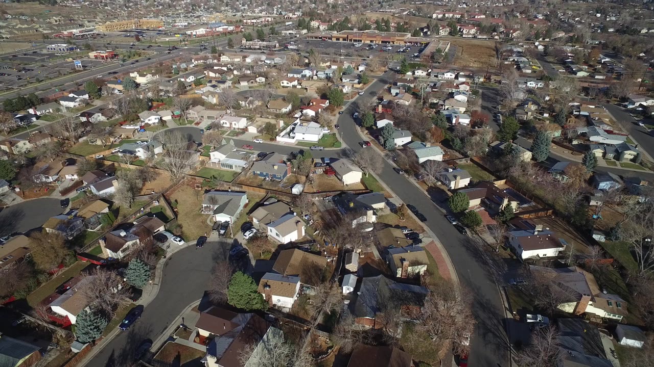 Aerial frame over Westminster, Colorado showing single-family homes, curved residential streets, parked cars, leafless trees, dry lawns, and no visible people.