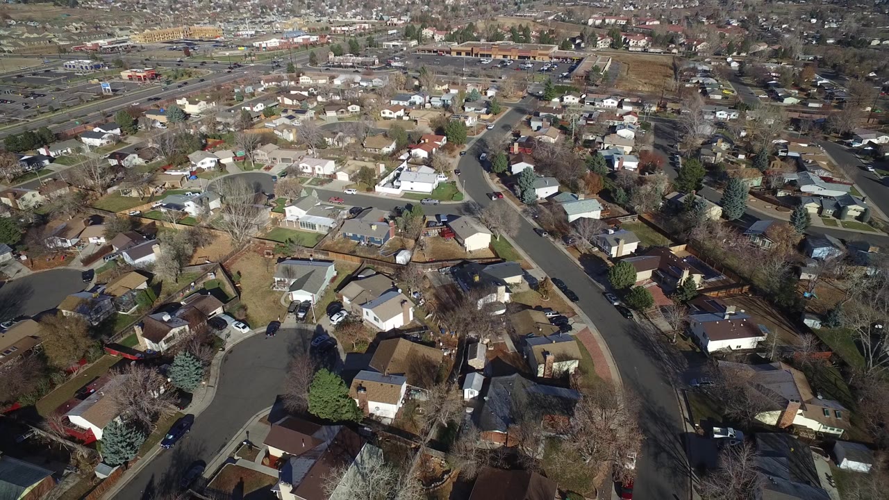High oblique aerial view in Westminster, Colorado with rooftops and backyards in the foreground, a looping street through the center, distant traffic roads, and no visible people.