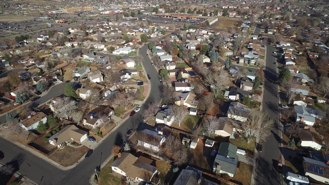 Aerial frame over Westminster, Colorado showing curved neighborhood roads, detached homes, bare trees, parked vehicles, and no visible people.