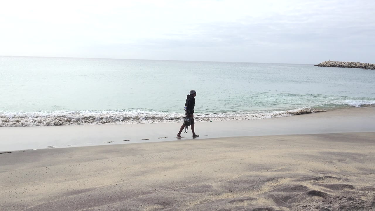 A man in dark clothing walks near the waterline on a San Jose del Cabo beach, with wet sand, pale surf, open ocean, and a distant rock breakwater visible.