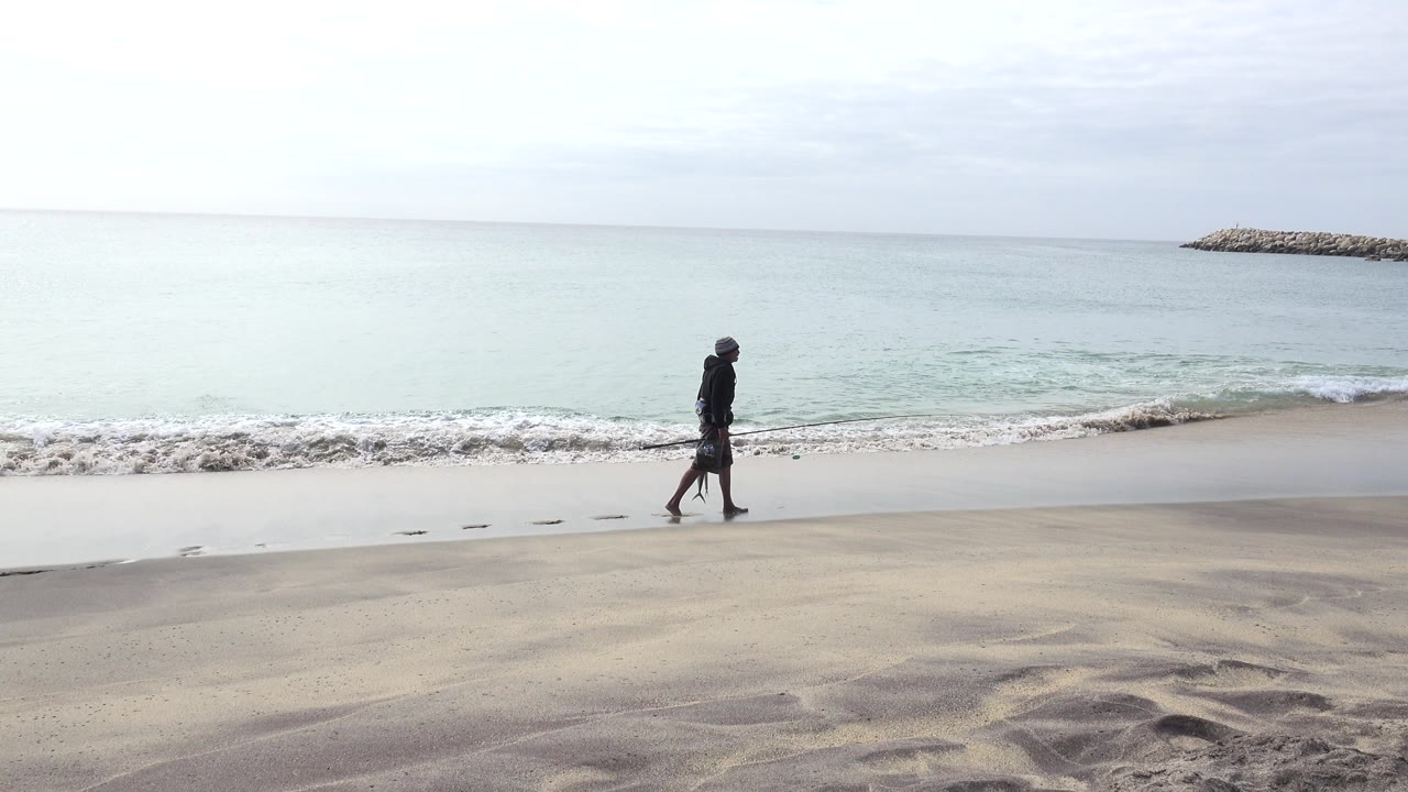 The man moves farther along the wet sand in San Jose del Cabo, with a low wave curling behind him and footprints trailing near the waterline.