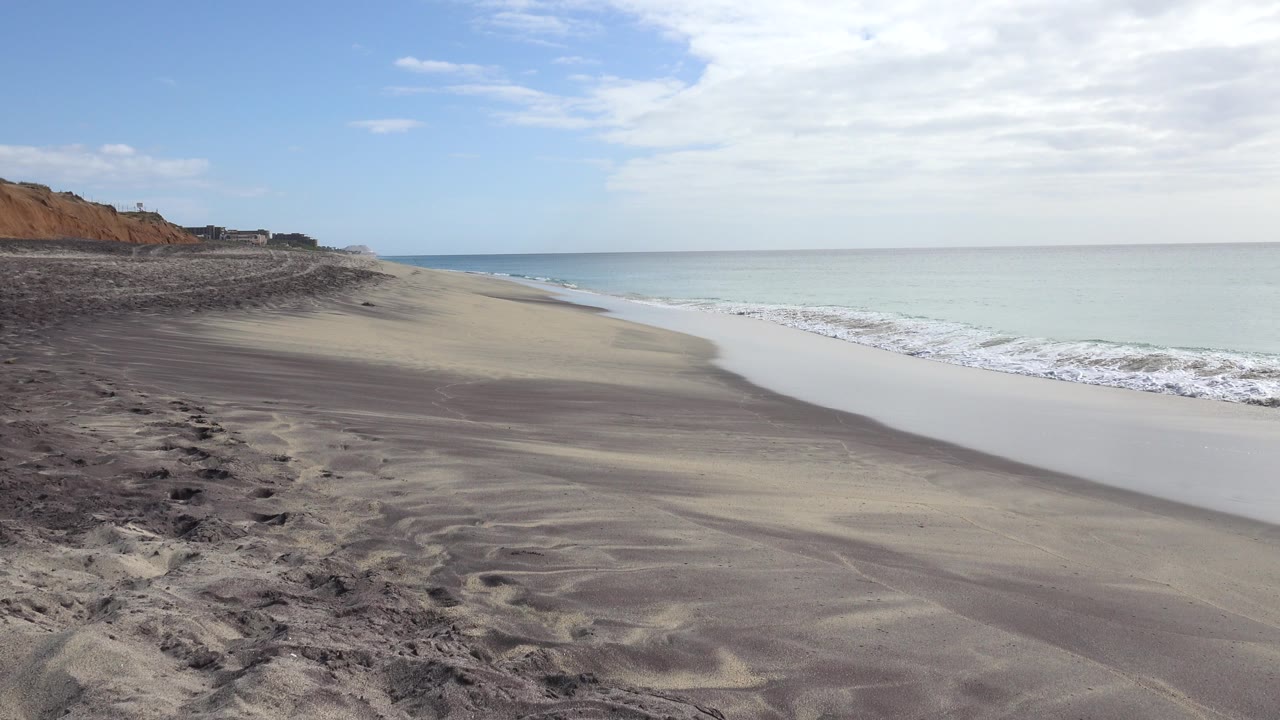 Empty beach with waves, sand, cliffs, ocean, and cloudy sky.
