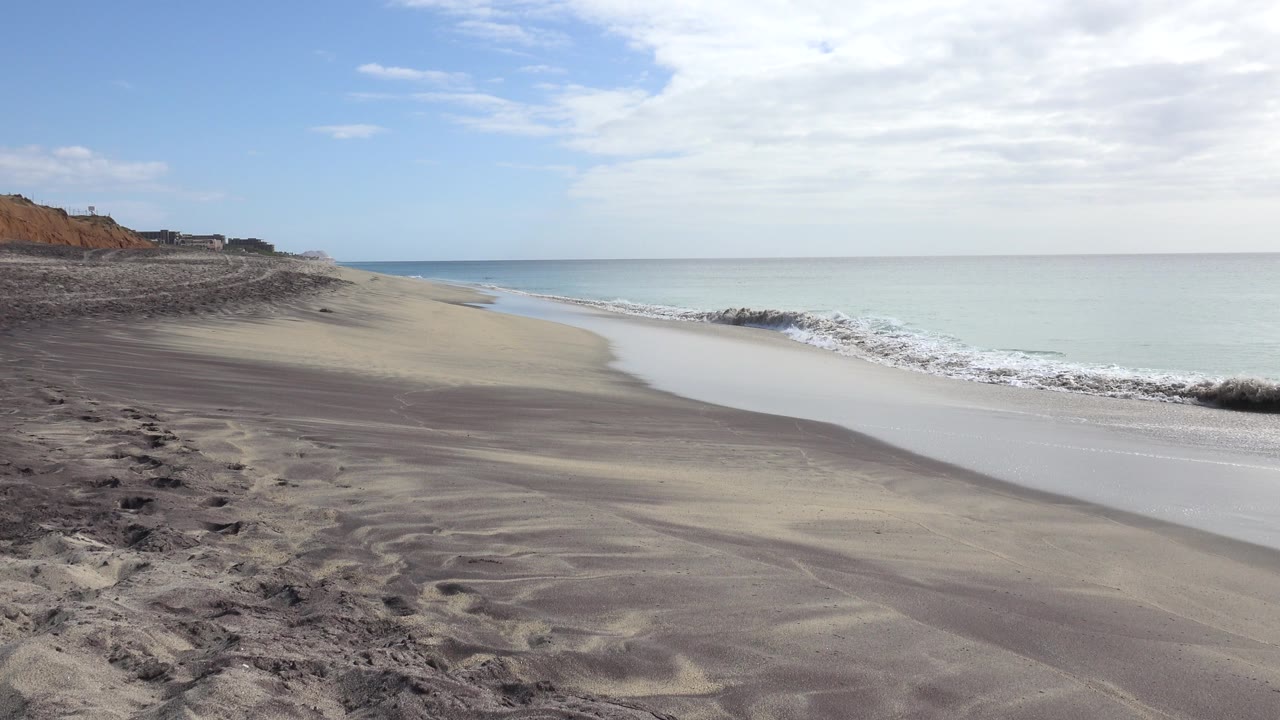 Empty sandy beach with dunes, surf, sea, and distant bluffs.