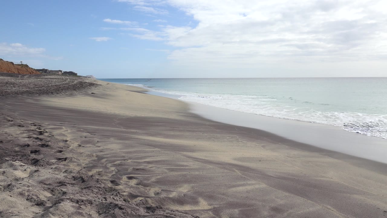 San Jose Del Cabo beach view at the start of the clip, with rippled sand in the foreground, surf washing along the right edge, distant bluffs to the left, cloudy sky above, and no visible people.