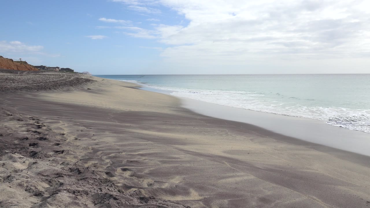 San Jose Del Cabo shoreline one second in, showing darker rough sand in the foreground, a smooth wet-sand strip, pale water on the right, reddish bluffs on the left, and no visible people.