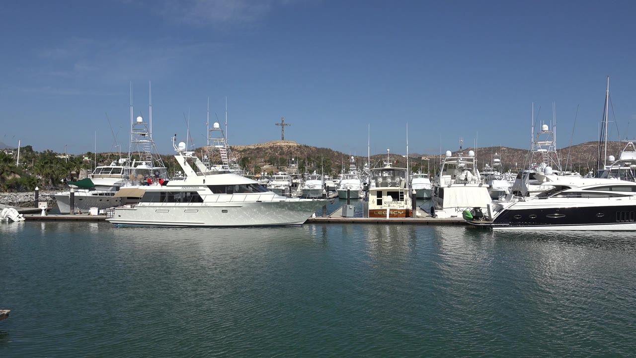 A steady San Jose del Cabo marina view shows white boats moored along the dock, rippled green-blue water in front, and low hills under bright daylight.
