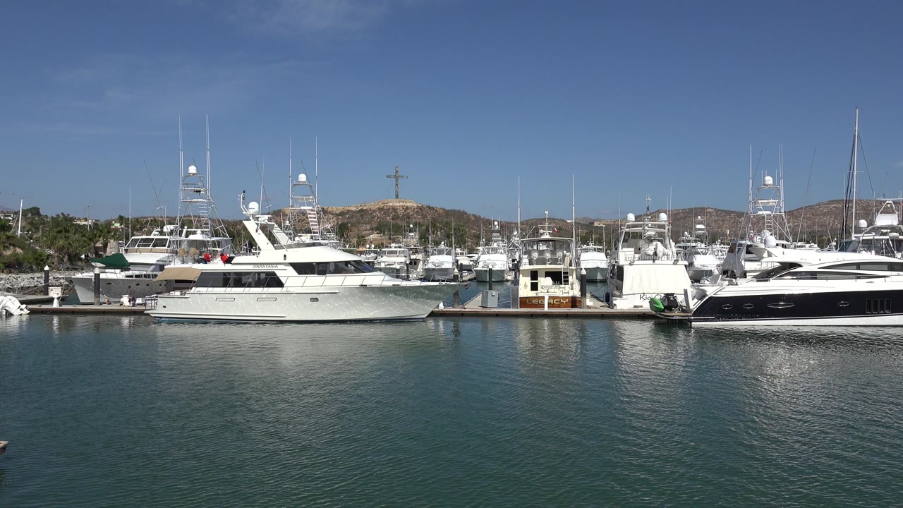 San Jose Del Cabo harbor frame with the large white yacht prominent at left, a dark-hulled yacht at right, rippled water in front, and no visible people.