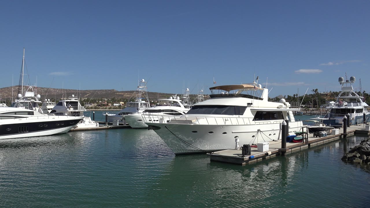 Opening frame of white yachts moored at a San Jose Del Cabo marina, with green-blue water in front, low hills behind, and no visible people.