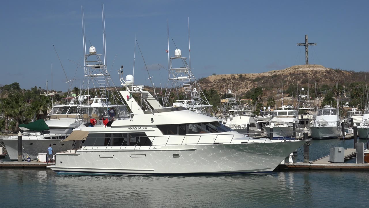 White yacht docked at a marina with people nearby and a hillside cross behind it.