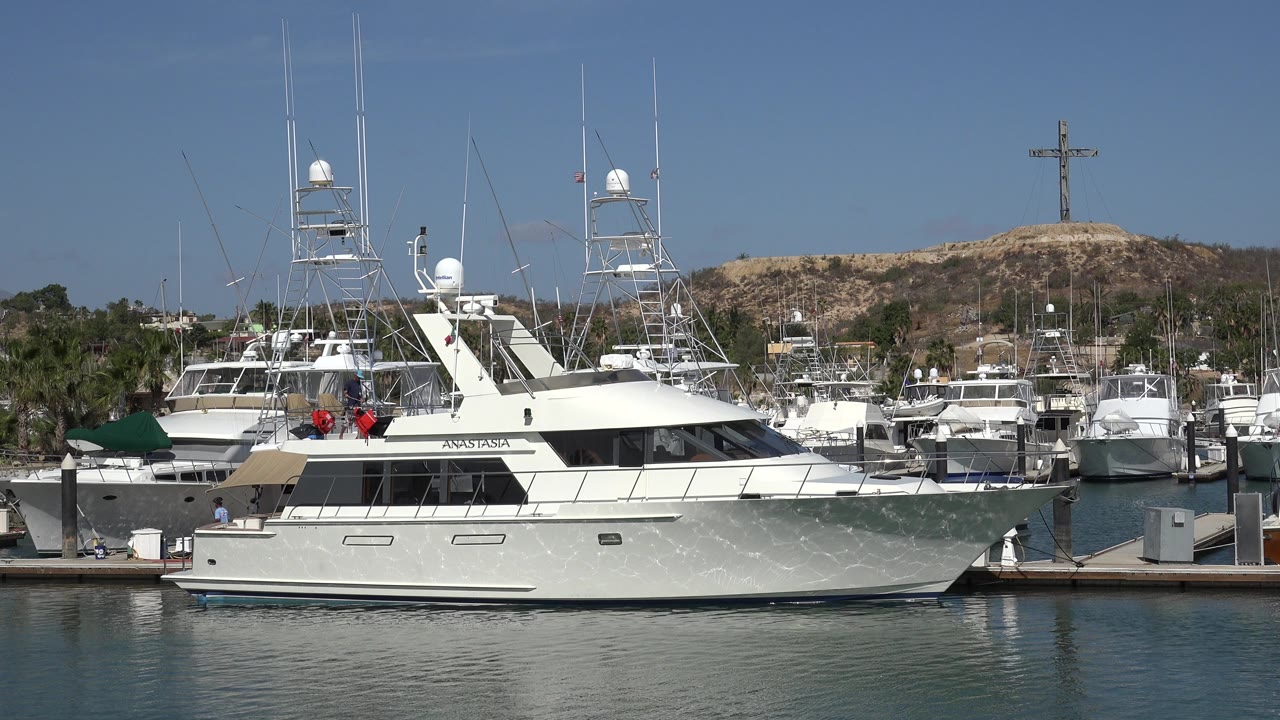 White yacht docked in a marina with boats and hillside behind it.