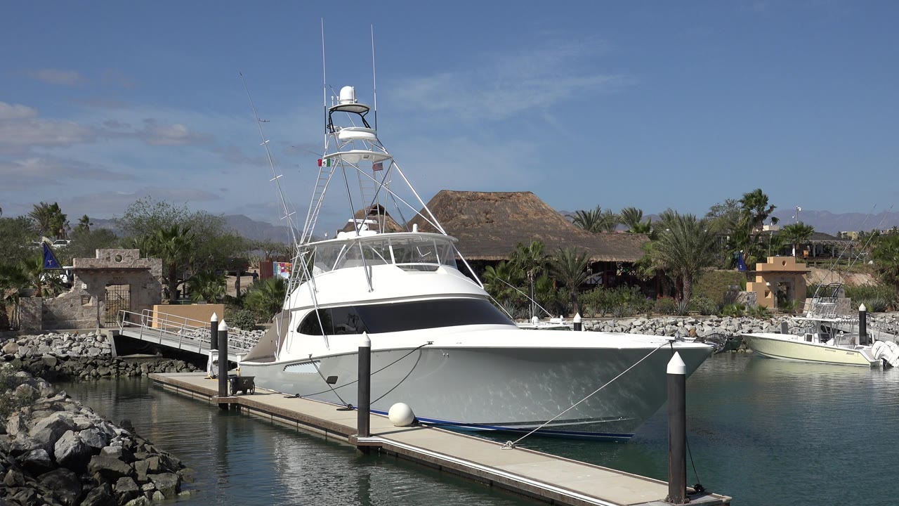 A large white yacht remains centered beside the floating jetty in San Jose Del Cabo, Mexico, with mooring lines crossing the bow, calm water, background palms, and no visible people.