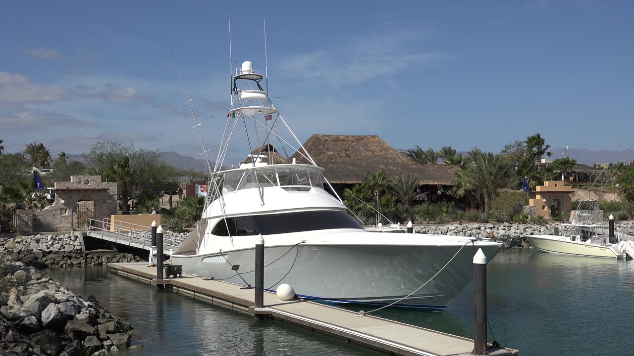 White sportfishing boat moored at a dock in San Jose del Cabo, Mexico, with rocks, calm water, palms, low buildings, distant hills, and no visible people.