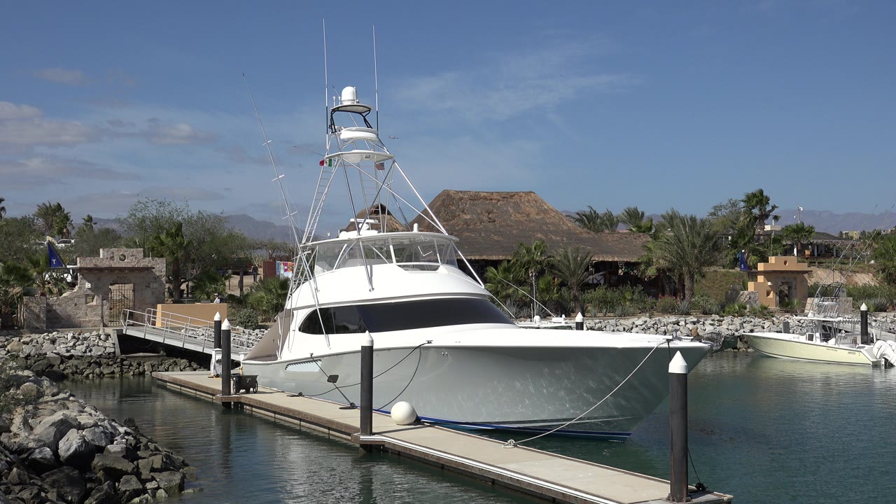 Large white boat remains tied beside the dock in San Jose del Cabo, Mexico, with the bow facing camera, still water below, waterfront buildings behind, and no visible people.