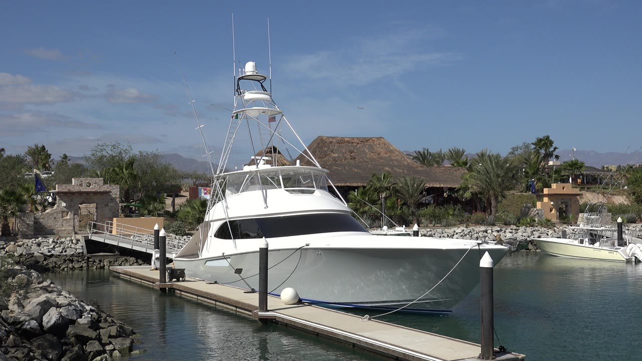 White sportfishing boat at a dock in San Jose del Cabo, Mexico, with rock breakwater in front, palms and low buildings behind, and tiny people possibly visible on the vessel.