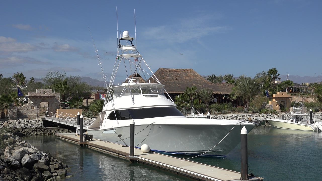 Moored white boat in San Jose del Cabo, Mexico, seen across still water with a foreground dock edge, black dock posts, palms, low buildings, and distant hills.