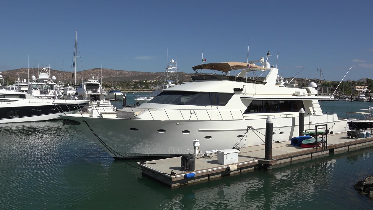 Large white yacht fills the center of the San Jose del Cabo marina view, with a dock corner in front, boats behind it, low hills, and no visible people.