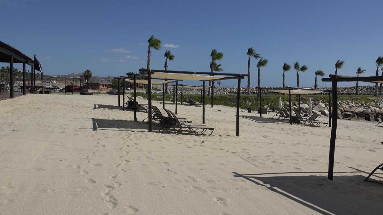 Opening frame of a San Jose del Cabo beach resort relaxation area with pale sand, empty lounge chairs, dark shade structures, palm trees, distant rocks, and no visible people.