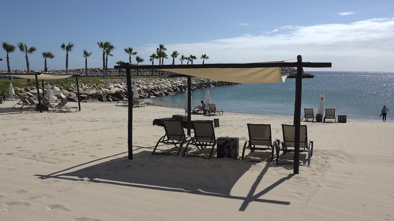White sand beach with shaded chairs, palms, rocks, blue water, and distant people.