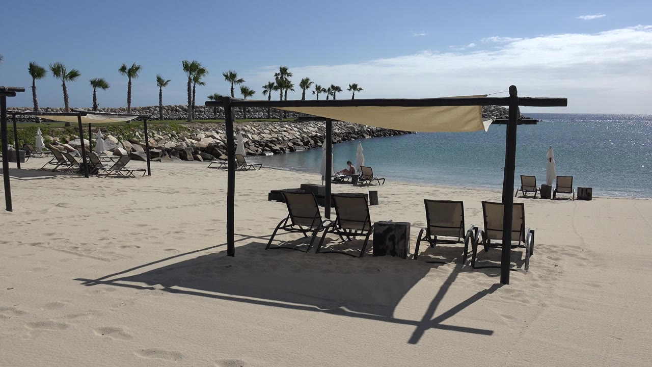 Cabana shade and lounge chairs on a sandy beach beside blue water.