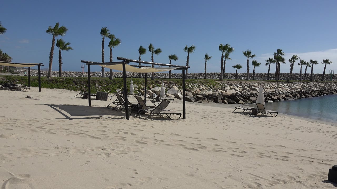 Quiet sandy beach with shade canopy, lounge chairs, palms, rocks, and calm water.