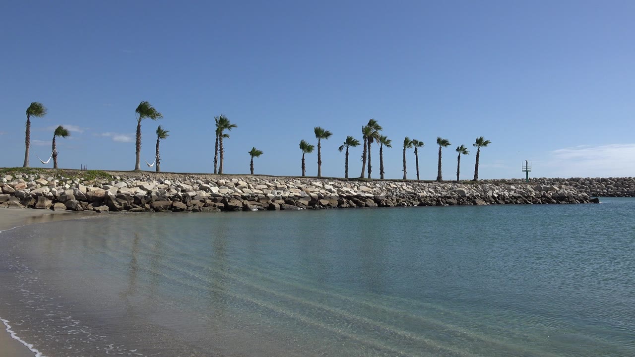 Opening frame of a San Jose del Cabo inlet with shallow blue-green water over sand, a rock barrier lined with palms, clear sky, and no visible people.