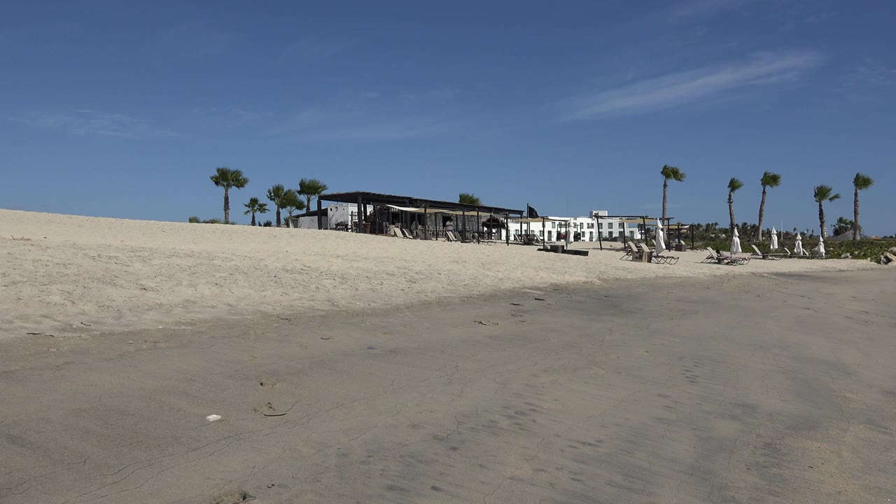 Opening frame of a San Jose del Cabo beach with wet sand in front, a low restaurant and hotel across the dunes, palm trees at right, and no visible people.