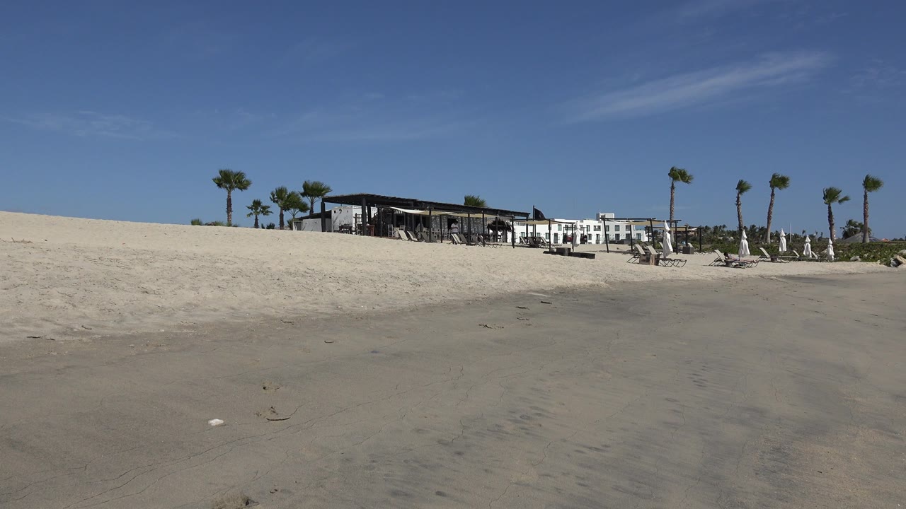 Second frame keeps the San Jose del Cabo beach centered on the hotel and restaurant, with pale sand, palm trees, beach chairs, umbrellas, and no visible people.