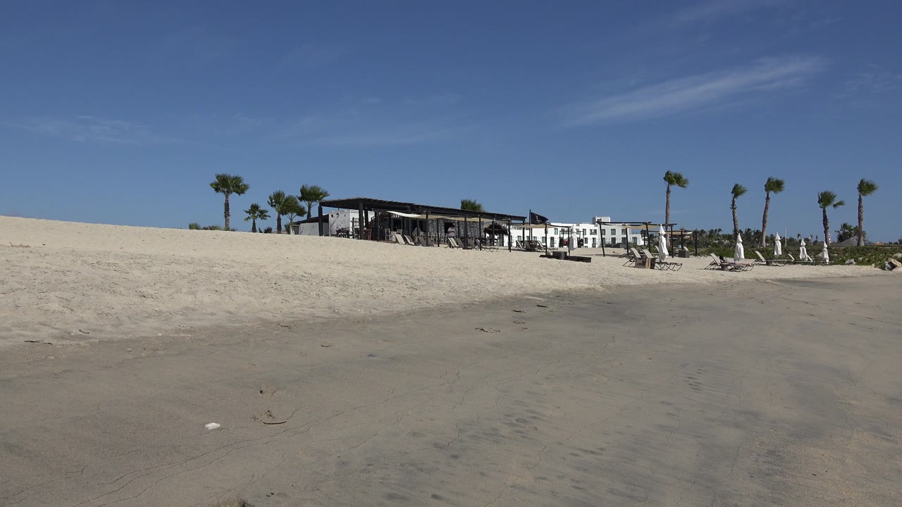 Beach sand leading toward a hotel, restaurant canopy, palm trees, and lounge area.