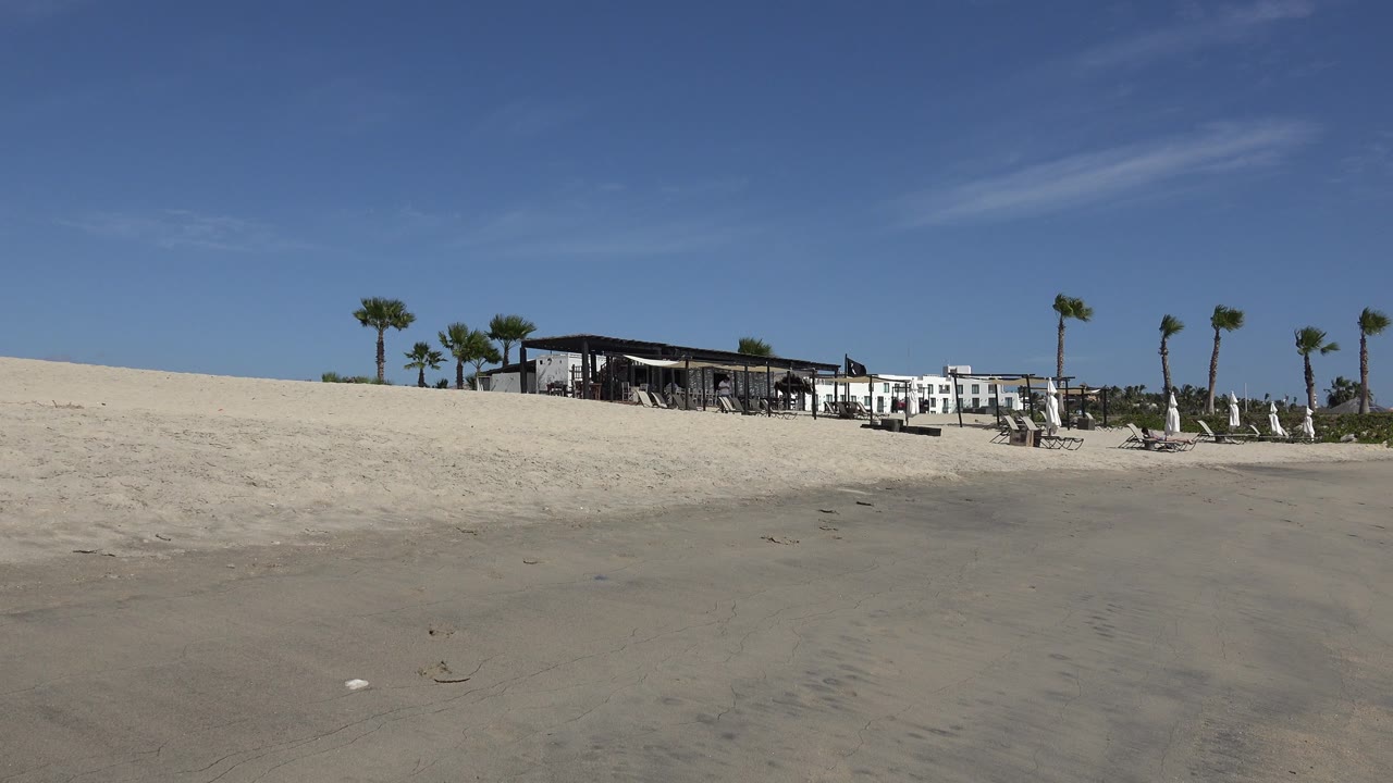 San Jose del Cabo beach frame one second in, with a broad sand foreground, the shaded building centered near the horizon, palm trees to both sides, and no visible people.