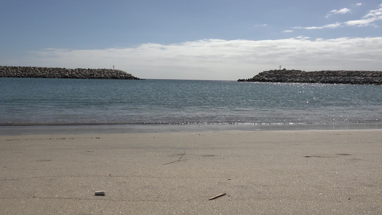 Opening frame of a San Jose del Cabo beach with sand in the foreground, calm blue water, rocky breakwaters on both sides, and no visible people.