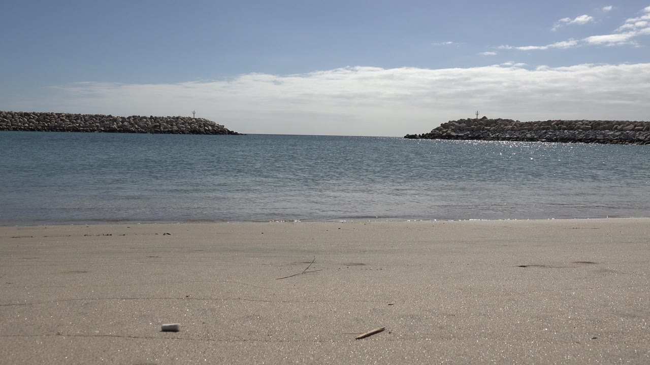 Empty sandy beach with calm sea and rocky breakwaters under blue sky.