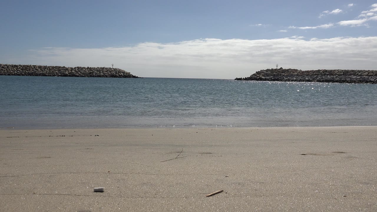 Opening frame of a San Jose Del Cabo beach with pale sand, calm water, rocky breakwaters on both sides, scattered clouds, and no visible people.