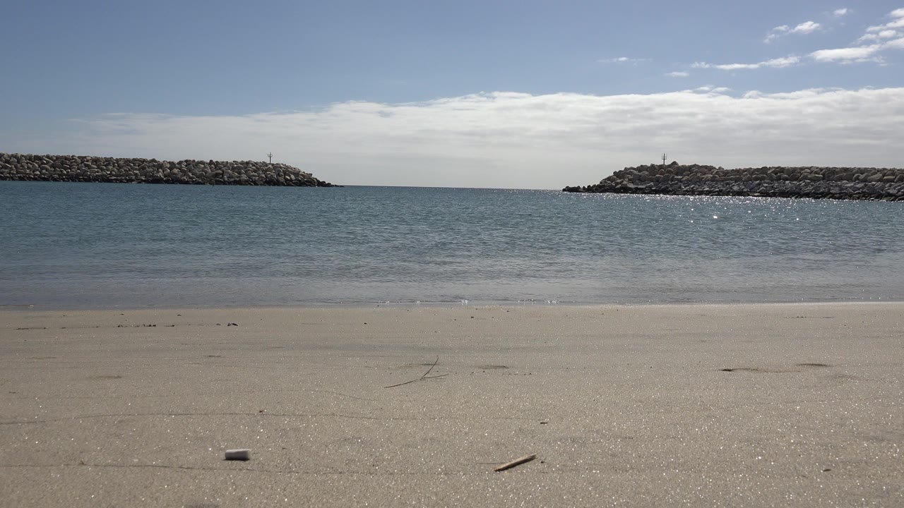 A San Jose Del Cabo shoreline view shows a thin wave line closer to the beach, rocky formations framing the water, bright sky, and no visible people.
