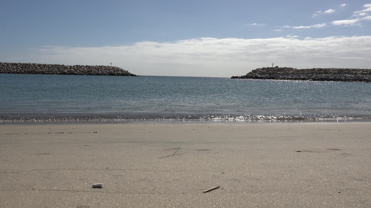 Empty sandy beach facing small waves and rocky breakwaters.