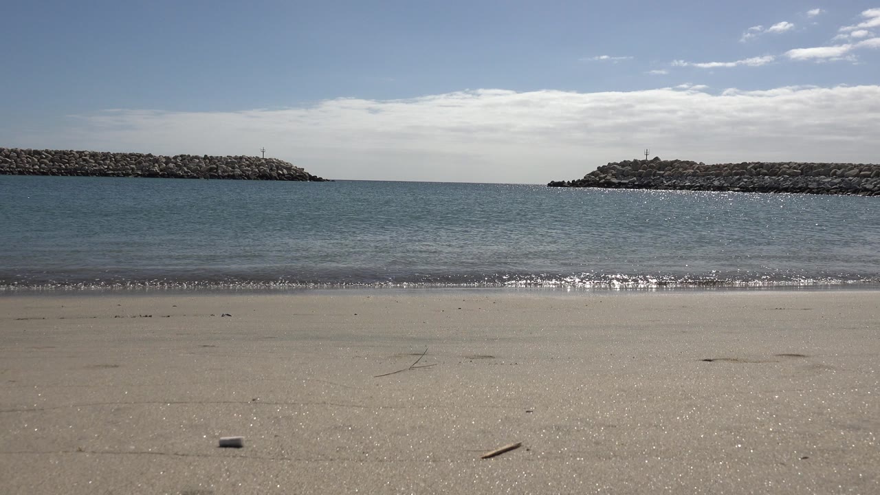 San Jose del Cabo shoreline with a thin surf line, glittering sunlight on the water, rocky barriers across the background, and no visible people.