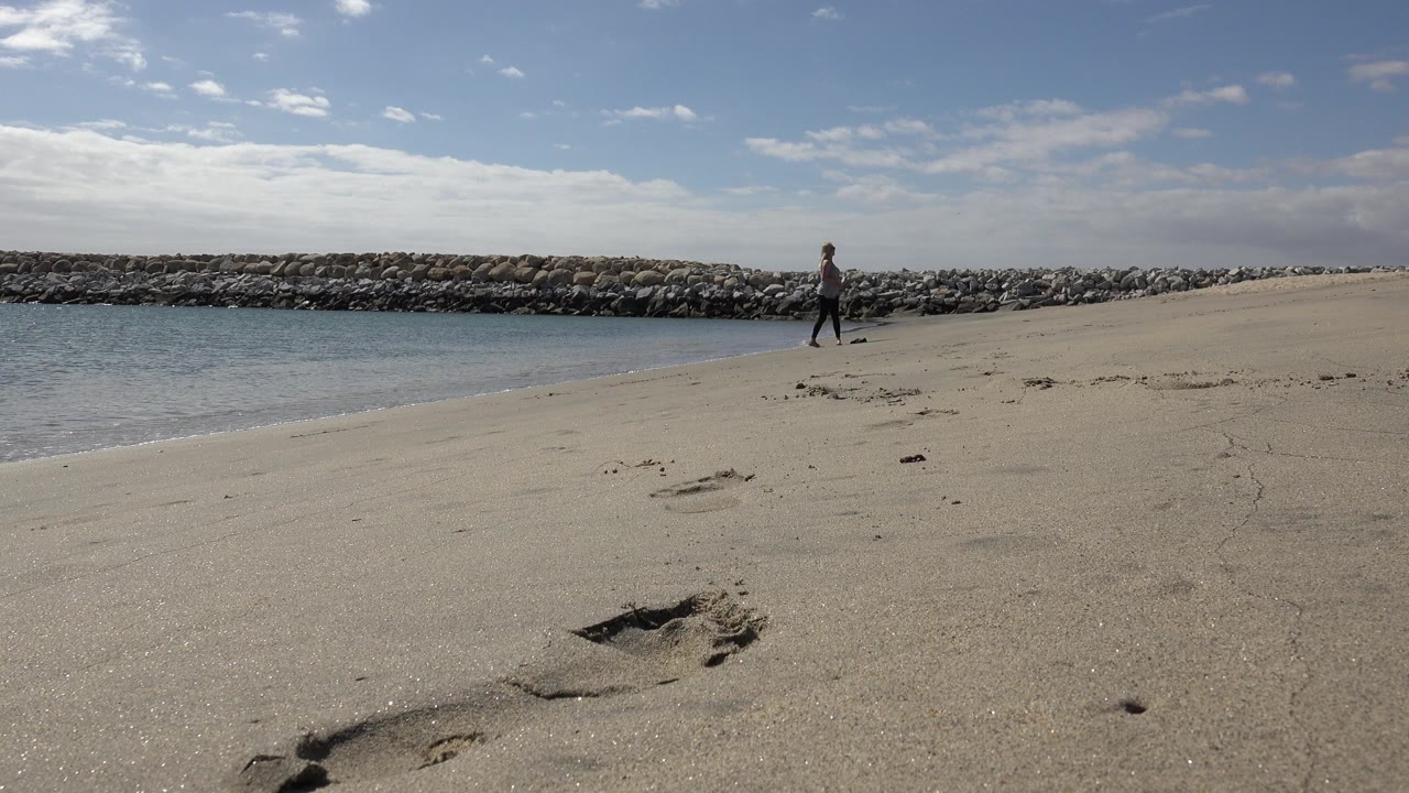 A wide view of San Jose del Cabo beach shows one person near the shoreline, calm water on the left, footprints in the sand, and a rock breakwater under scattered clouds.