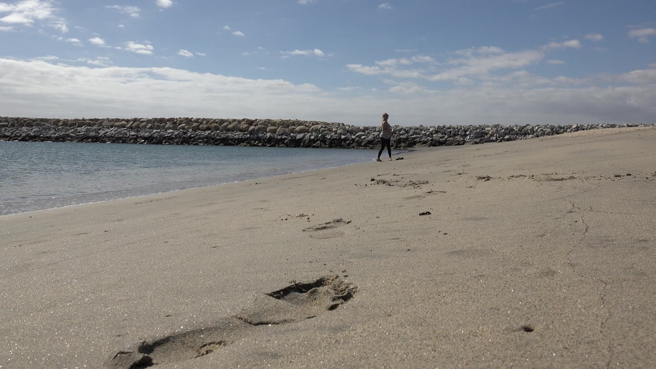 At 0:01, the person remains near the waterline on the San Jose del Cabo beach while the foreground footprints and long rock barrier stay prominent in the wide composition.