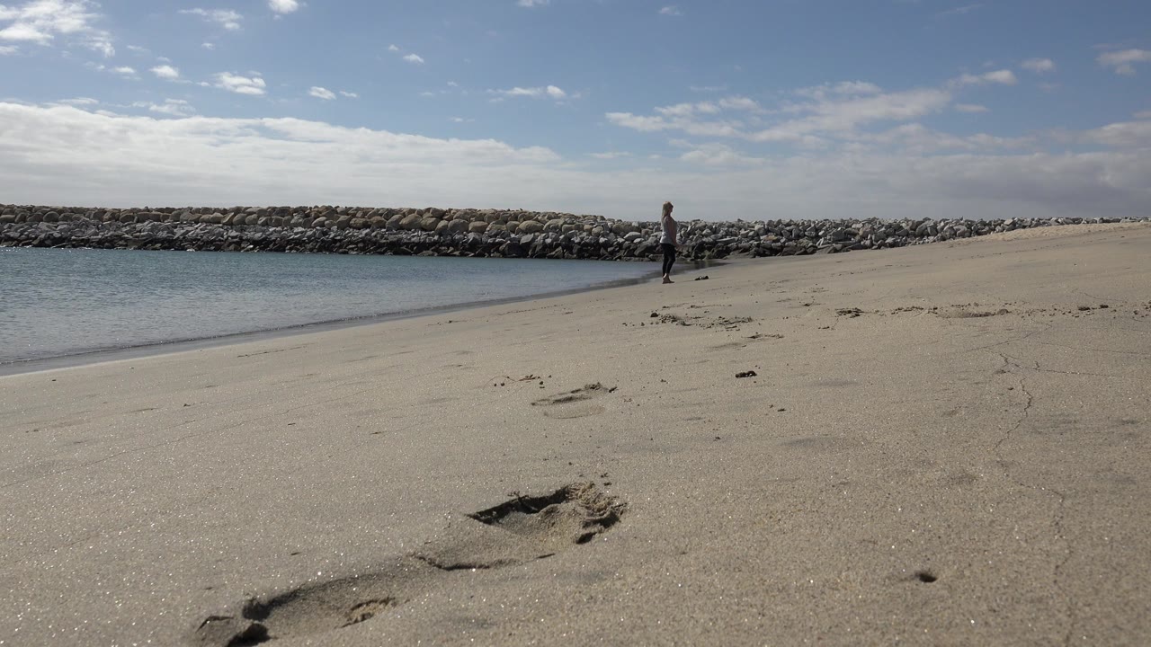 Sandy beach with footprints, water, rock jetty, and one distant person.