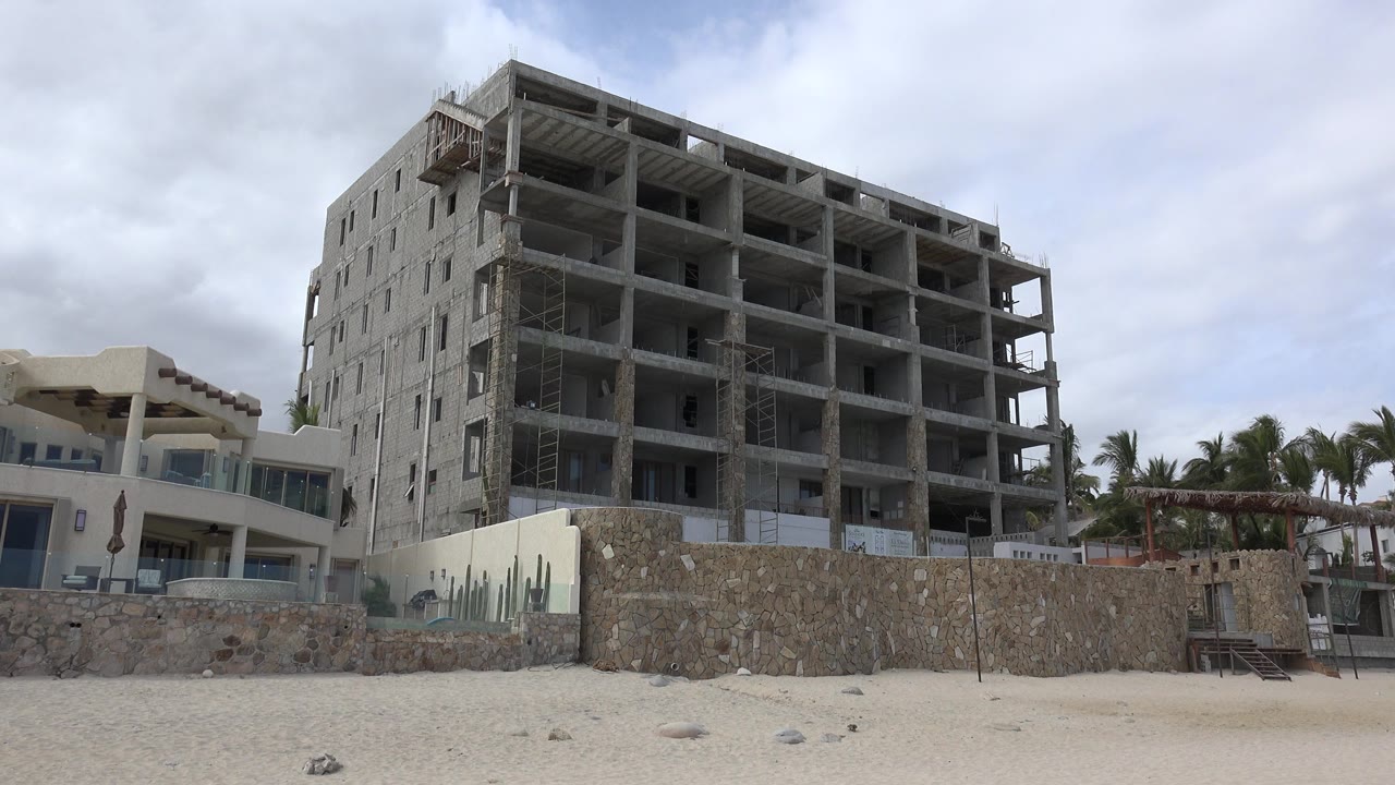 Wide view in San Jose del Cabo, Mexico of an unfinished concrete building behind a stone wall, with sandy foreground, cloudy sky, palm trees, and no visible people.