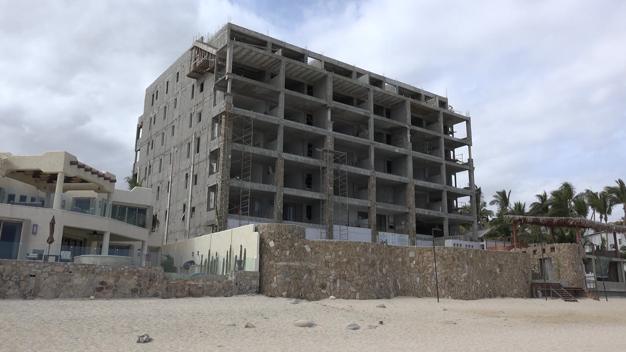 Opening frame of an unfinished concrete building in San Jose del Cabo, Mexico, seen behind a stone wall with sand in front and no visible people.