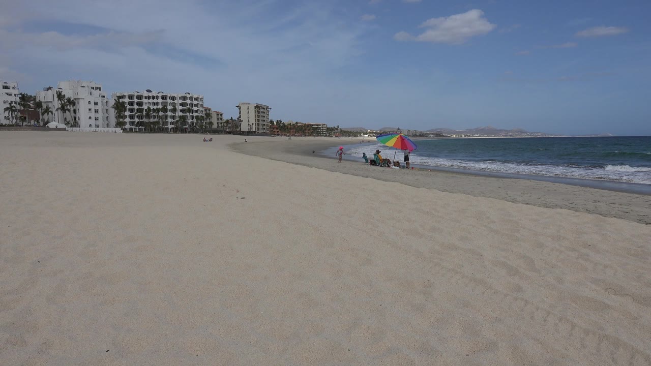 San Jose del Cabo shoreline view with the umbrella group slightly right of center, broad sand in the foreground, low surf along the waterline, and high-rise buildings in the left background.